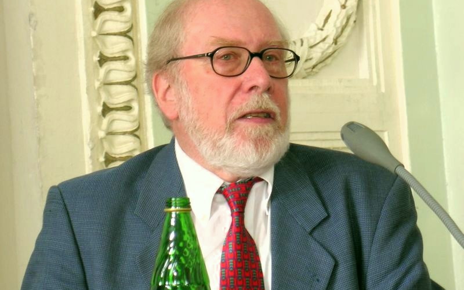 Bearded man in glasses and suit jacket speaking into a microphone at a podium